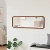 A long, rectangular mirror with a brown frame hangs on a white wall above a black bench with a checkered pillow. A potted plant with large green leaves is in the foreground. Voir les images du produit