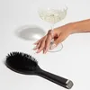 A hand with manicured nails rests near a champagne coupe filled with a light-colored beverage, next to a black hairbrush on a white background. Produktbild-Vorschau