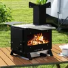 A camping stove with a fire burning inside, set up on a wooden table outdoors with grass and a tent in the background. Produktbild-Vorschau