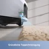 A robotic vacuum cleaner is shown transitioning from a hard floor to a carpet, with a blue light indicating its sensor system. Produktbild-Vorschau