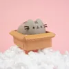 A Pusheen figurine sits in a cardboard box, surrounded by white packing peanuts, against a pink background. Produktbild-Vorschau