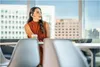 A woman wearing a headset is sitting at a table in a modern office setting with large windows overlooking a city. Produktbild-Vorschau