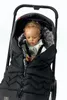 A baby is sitting in a stroller, wrapped in a black footmuff with a fur-lined hood. Náhled obrázku produktu