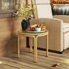 A rattan side table with a glass top sits on a wooden deck, holding a vase of flowers and a plate of fruit. A wicker chair is visible in the background. Podgląd zdjęcia produktu