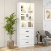 A white display cabinet with glass doors and drawers, illuminated by LED lights, stands next to a potted plant in a room. Podgląd zdjęcia produktu