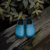 A pair of blue clogs rests on a weathered wooden surface, with lush green foliage in the background. Produktbild-Vorschau