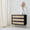 A black chest of drawers with rattan fronts and wooden legs, placed next to a cream-colored curtain. A vase with dried grass is on top of the chest. Produktbild-Vorschau