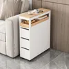 A narrow white cabinet with wooden accents and drawers sits next to a sofa, with a magazine and cup on top. Produktbild-Vorschau