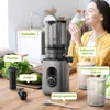 A woman is drinking juice from a glass next to a slow juicer and its accessories on a wooden table. Produktbild-Vorschau