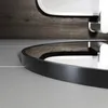 A close-up view of the edge of a round black mirror with a brushed aluminum frame, resting on a light gray surface. The background shows a dark wood-paneled wall and a reflection of the mirror and wall. Náhľad obrázku produktu