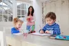 A woman is standing behind two children who are sitting at a table and drawing. Anteprima dell’immagine prodotto