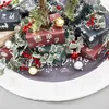 A collection of Christmas gifts wrapped in festive paper, adorned with holly and berries, arranged around the base of a Christmas tree. Produktbild-Vorschau