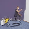 A man is painting a wall with a spray gun, with a paint sprayer machine and bucket nearby. Náhled obrázku produktu