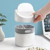 A hand is shown placing the lid on a small white trash can with a gray liner, on a desk next to a laptop and a plant. Produktbild-Vorschau
