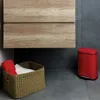 A red trash can and a woven basket with towels are placed on a tiled floor next to a wooden cabinet. Produktbild-Vorschau