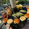 A large, round metal grill surface with various foods being cooked on it, including burgers, sausages, chicken, asparagus wrapped in bacon, and corn on the cob. There are also burger buns on the grill. The grill is part of a larger setup, possibly a fire p Produktbild-Vorschau