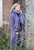 A woman wearing a long purple knitted cardigan and scarf stands in front of a brick wall with dry grass in the foreground. Produktbild-Vorschau