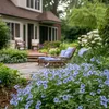 A lush garden scene with blue flowers in the foreground, a patio with lounge chairs in the midground, and a house in the background. Produktbild-Vorschau
