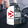 A Dymatize Super Mass Gainer product is shown in a gym setting with a person's legs and a barbell in the background. Produktbild-Vorschau