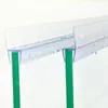 A close-up shot of a clear plastic shower door seal with water droplets on it, attached to two green glass panels. Podgląd zdjęcia produktu