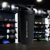A black Smartshake shaker bottle is displayed in a gym setting, with various fitness equipment like medicine balls and kettlebells on shelves in the background. Produktbild-Vorschau
