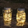 Two solar-powered mason jar lights with warm white LEDs are placed on a wet, rocky surface at dusk, with raindrops visible in the air. Produktbild-Vorschau