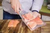 A person is holding a vacuum bag filled with salmon fillets over a wooden cutting board. Produktbild-Vorschau