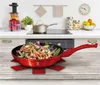 A red frying pan with food is placed on a wooden table in a kitchen setting. The background shows kitchen appliances and countertops. Náhled obrázku produktu