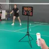 A digital scoreboard is set up on a tripod on a green badminton court, with a person playing badminton in the background. Podgląd zdjęcia produktu