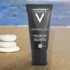 A tube of Vichy Dermablend foundation is shown on a sandy beach with smooth white stones in the foreground and the ocean in the background. Podgląd zdjęcia produktu