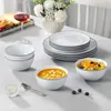 A table setting with a set of white porcelain dishes, including plates and bowls, with a black rim line. There is also a glass of red wine, a tray of pastries, a vase of flowers, and some fruit. Produktbild-Vorschau