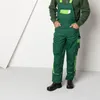 A person wearing green overalls and a green sweatshirt stands in front of a white and gray background. Produktbild-Vorschau