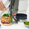 A person is adding lime to a layered lunch bowl. A separate container lid and a bowl of edamame are also visible. Anteprima dell’immagine prodotto