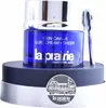 A jar of La Prairie Skin Caviar Luxe Cream Sheer is displayed on a dark blue and silver stand, with a silver spatula resting beside it. A circular emblem with a building illustration and the La Prairie logo is attached to the stand. Voir les images du produit
