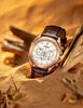 A rose gold Citizen watch with a brown leather strap is partially buried in sand, surrounded by driftwood. Anteprima dell’immagine prodotto