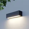 A modern, rectangular LED wall light mounted on a textured wall, with green leaves visible in the upper left corner. Produktbild-Vorschau