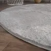 A close-up view of a round, fluffy gray rug on a wooden floor. A white basket is visible in the background. Voir les images du produit