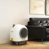 A white and gray automatic cat litter box is placed on a wooden floor next to a black leather sofa in a living room. Produktbild-Vorschau