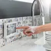 A person with soapy hands is pressing a button on a white device mounted on a tiled wall next to a kitchen sink with running water. Podgląd zdjęcia produktu