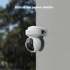 A white security camera is mounted on a gray wall, with rain falling on both the camera and the wall. The background shows blurred trees. Produktbild-Vorschau