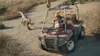 A person in a yellow hard hat drives an ATV through a dry, brushy landscape, with two raptors nearby. Náhľad obrázku produktu