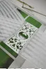 A close-up view of a white decorative panel with intricate cutouts, bordered by a silver ribbon, placed on a striped white sheer curtain. The background shows a blurred green outdoor scene through a window. Náhľad obrázku produktu