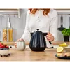 A woman is in a kitchen, holding a mug next to a black electric kettle. There are fruits and other kitchen items on the counter. Produktbild-Vorschau