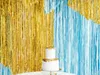 A two-tiered white cake with gold trim sits in front of a backdrop of gold and blue metallic fringe curtains. Produktbild-Vorschau