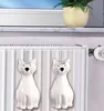 Two white ceramic cat-shaped humidifiers hang on a radiator, with a potted plant visible on a windowsill in the background. Podgląd zdjęcia produktu