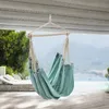A teal hammock chair is suspended from a wooden beam on a porch, with a blurred background of trees, water, and mountains.