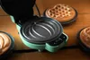 A close-up view of an open mini waffle maker with a green exterior and black interior, showing the waffle plates. Several finished waffles are visible in the background and foreground. Náhled obrázku produktu