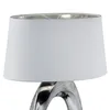 A close-up view of a table lamp with a white fabric shade and a silver-colored ceramic base. Náhled obrázku produktu