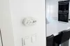 A white carbon monoxide detector is mounted on a white wall next to a light switch. In the background, a modern kitchen with white cabinets and stainless steel appliances is visible. Podgląd zdjęcia produktu