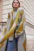A woman wearing a long, textured cardigan with a geometric pattern of gray and mustard yellow, standing outdoors in front of a stone building. Produktbild-Vorschau